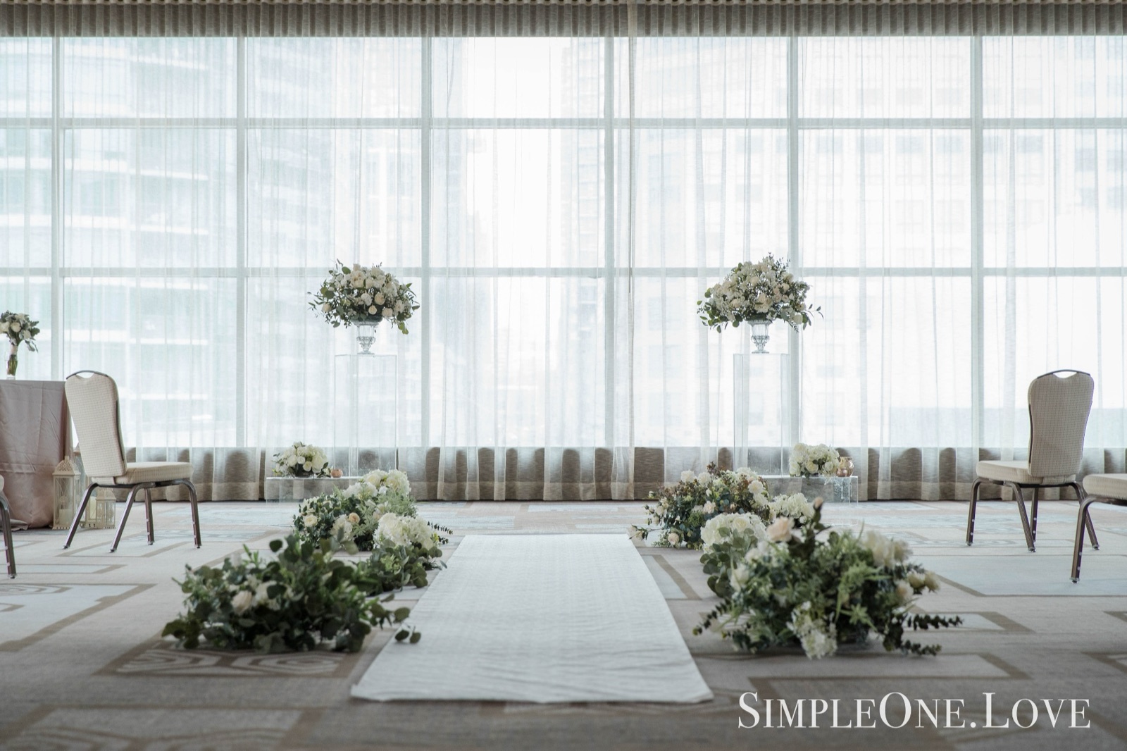 A hotel hall dressed for a ceremony, with a white aisle and tall windows.