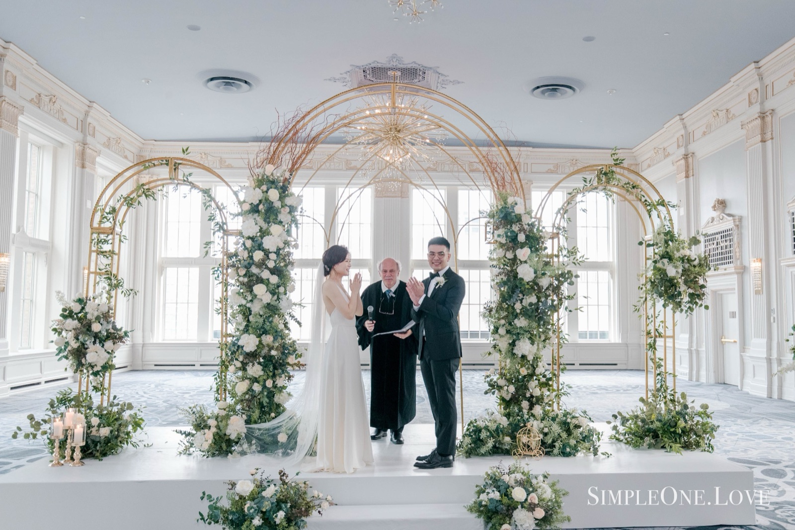 A couple and their officiant under a gilded arch in a bright hall.