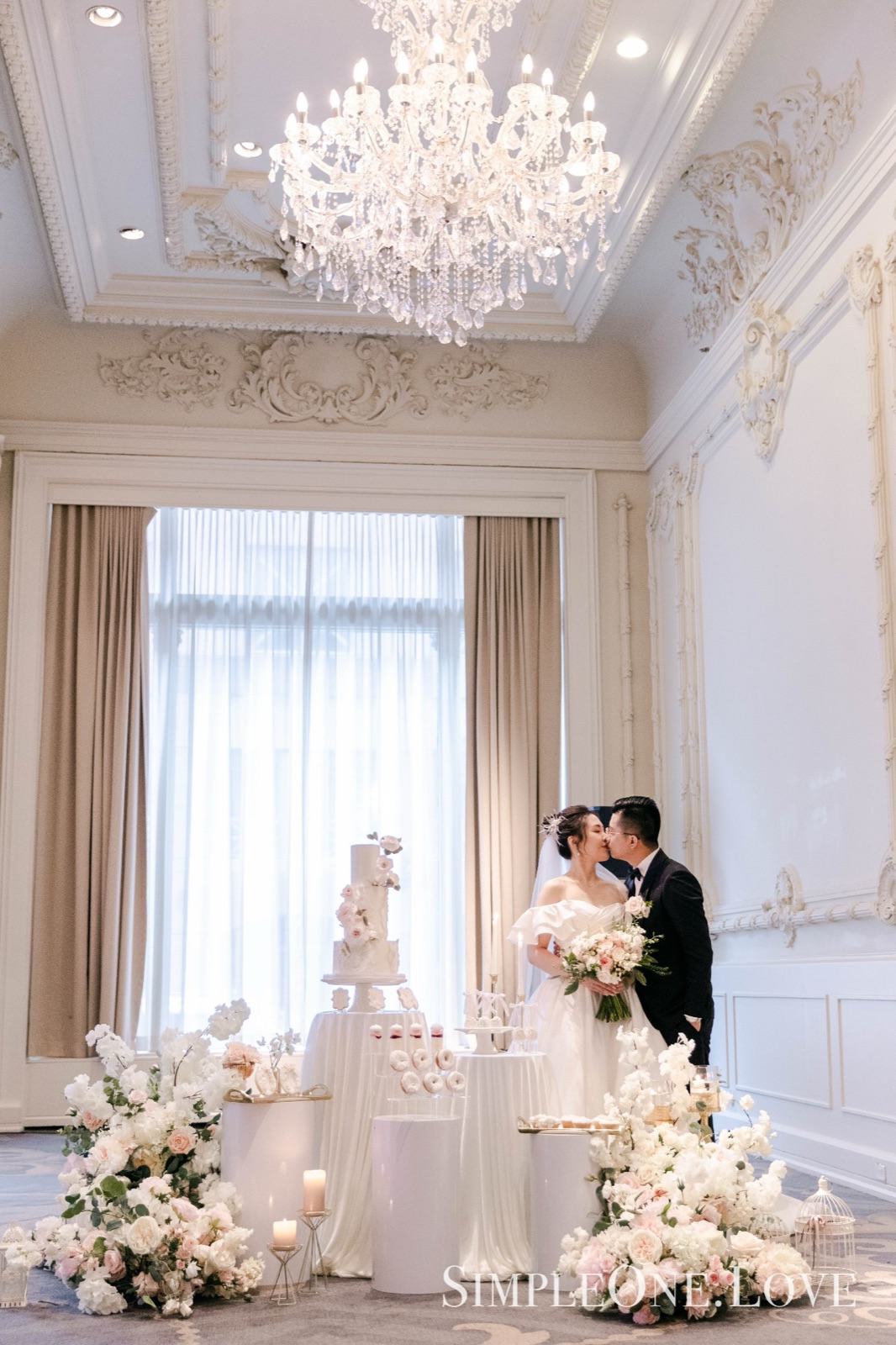 A couple beside their cake in a chandelier-lit room.