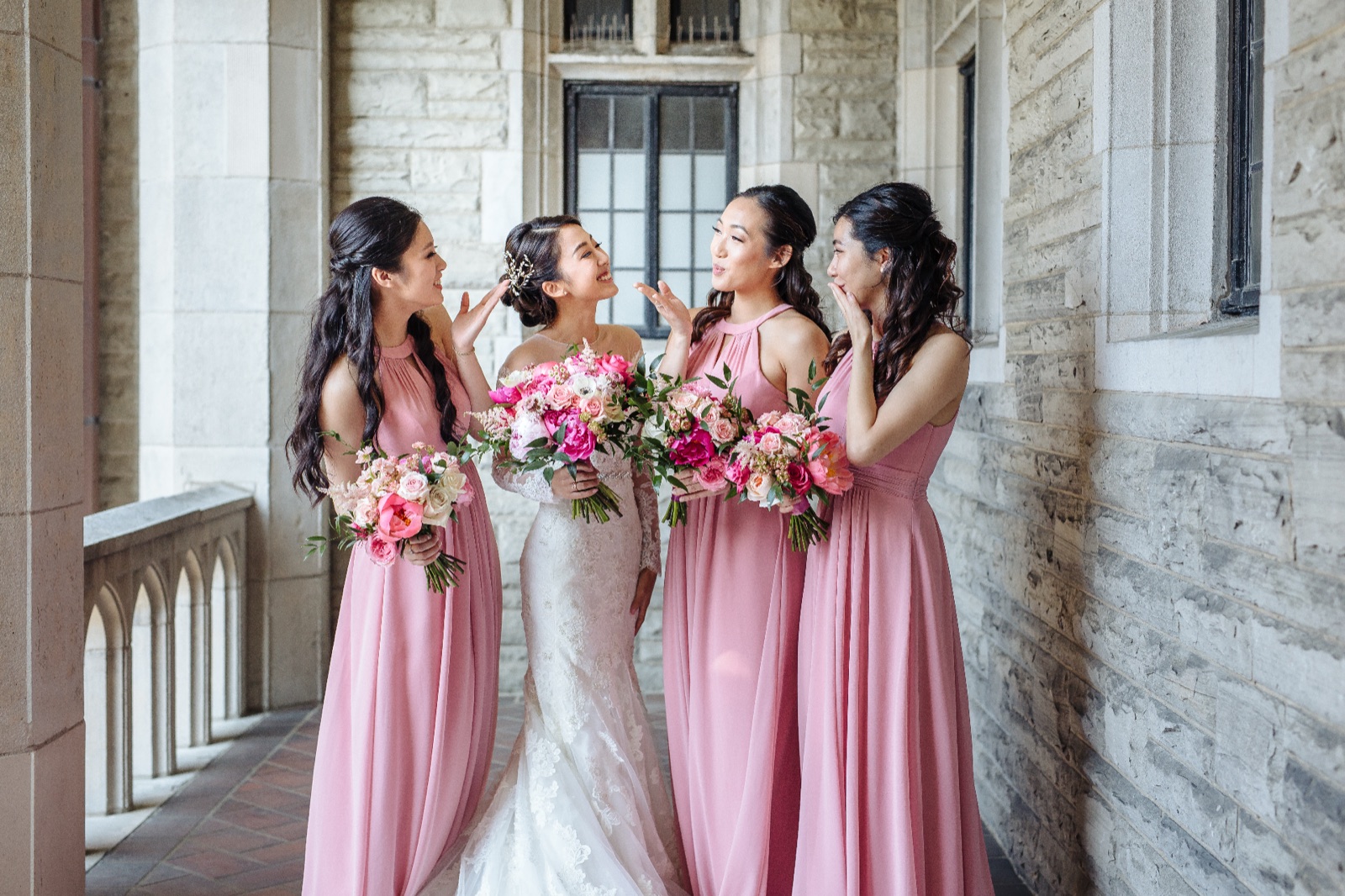 A bride and her bridesmaids share a laugh under a stone arcade before the ceremony.